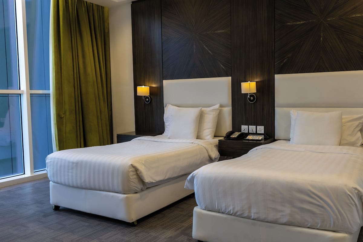 A hotel room with two beds, a large window with curtains, and wall-mounted lamps.