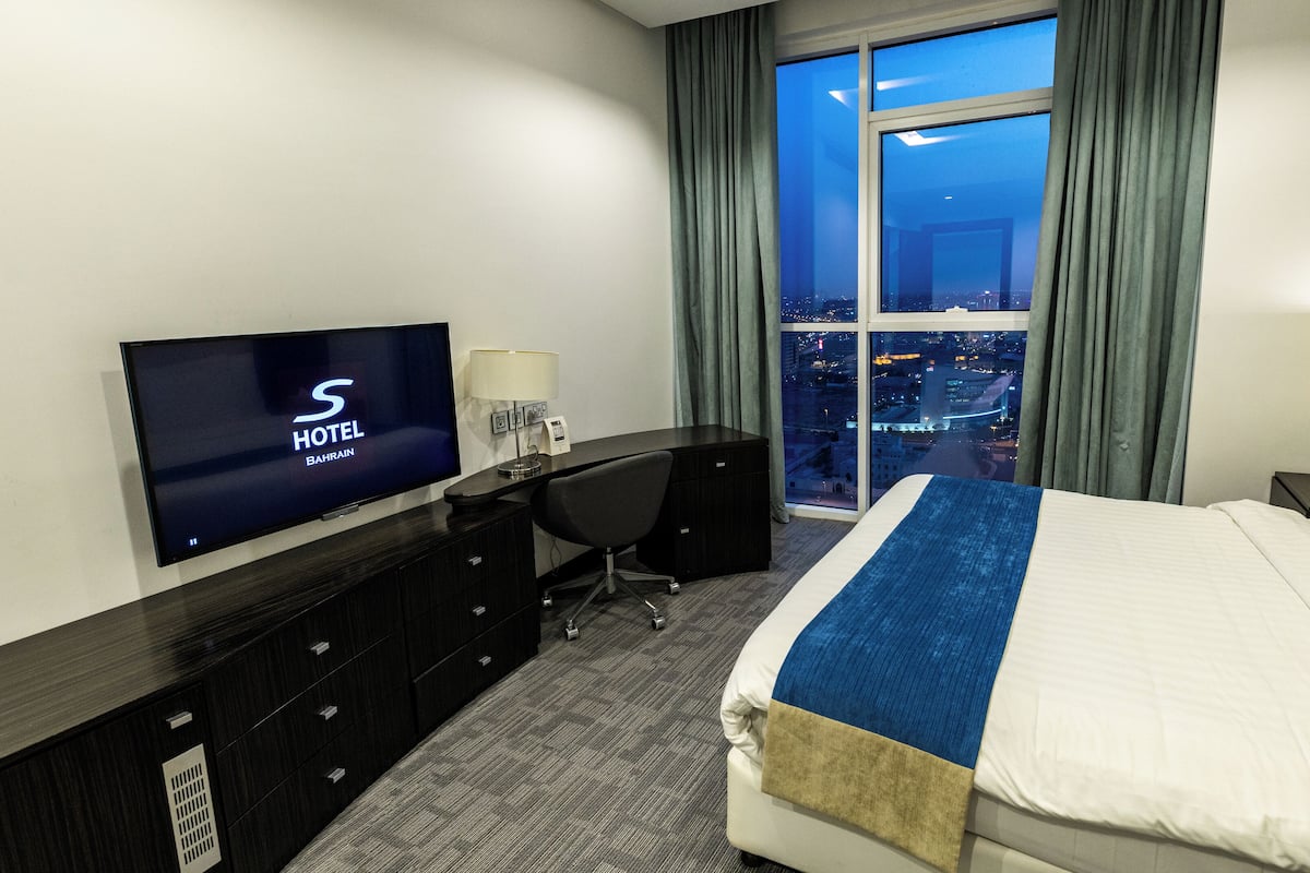 A hotel room with a bed, a desk, a TV displaying 'S HOTEL BAHRAIN', a city view, and a night sky visible through the window.