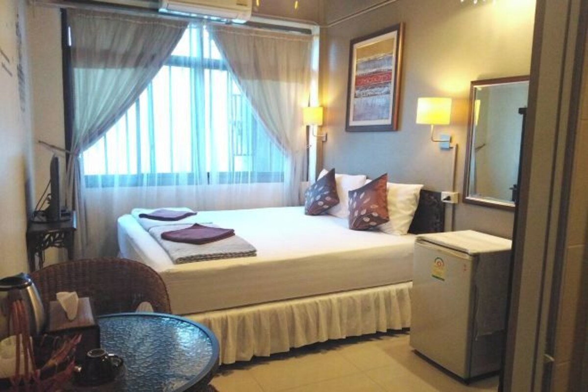 Une chambre d’hôtel avec un lit, un petit bureau, une chaise, une table, un miroir et une fenêtre avec des rideaux.