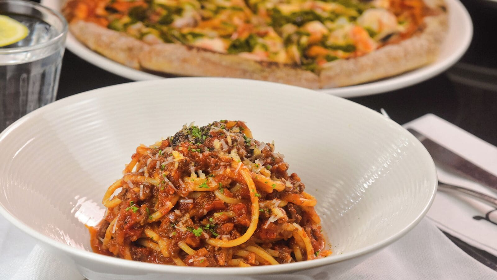 En skål med spaghetti med en tomatsås, garnerad med örter, och en pizza i bakgrunden.