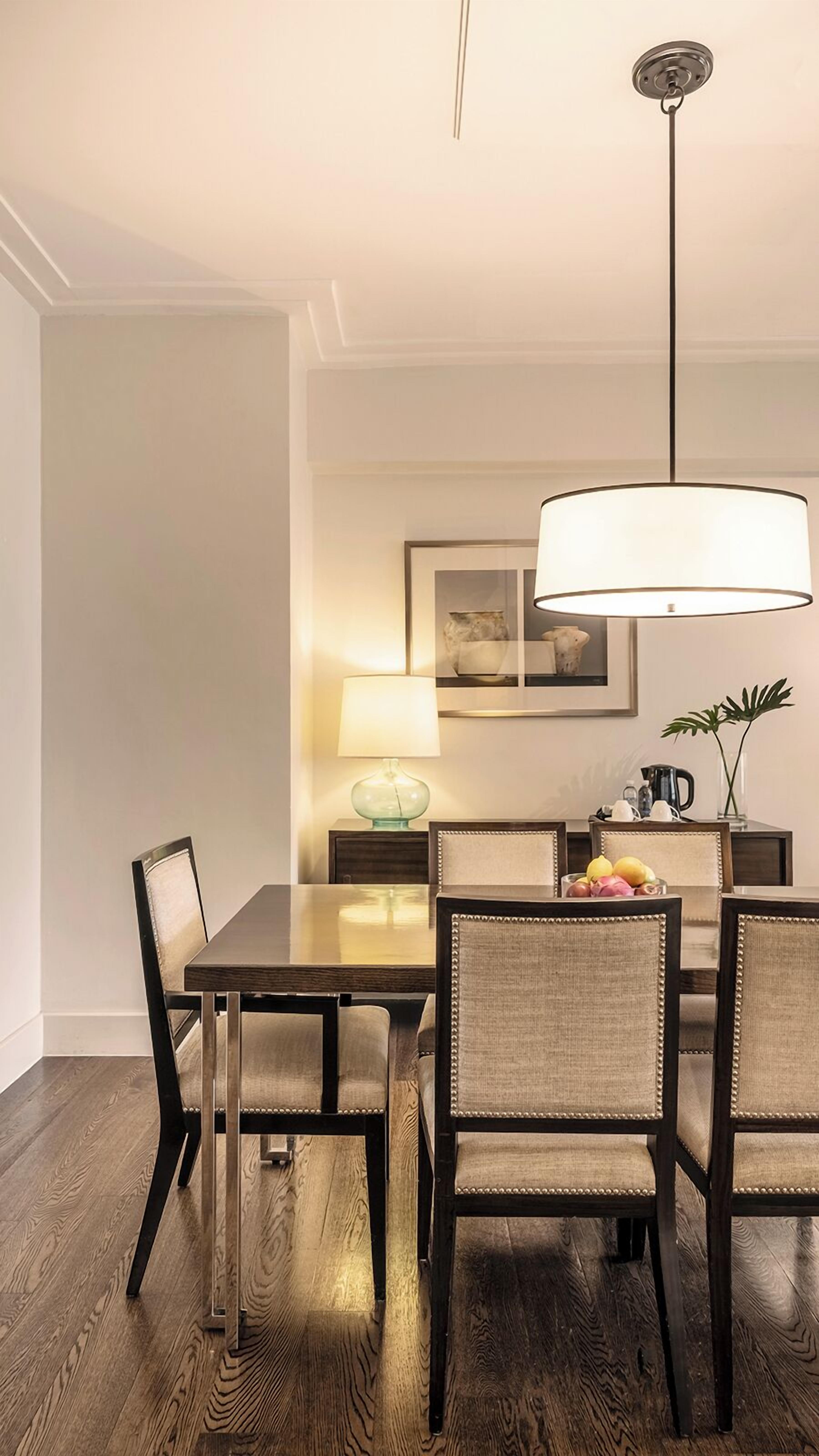 A dining area with a wooden table, four upholstered chairs, a hanging light fixture, a side table with a lamp, and a decorative vase.