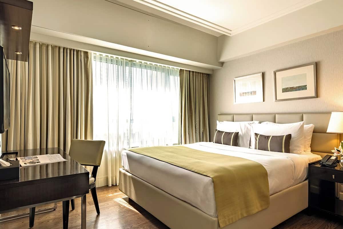 Habitación de hotel con cama, escritorio, silla, mesita de noche con lámpara y ventana con cortinas.