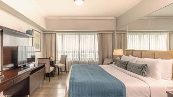 A hotel room with a bed, a desk with a television, a chair, and a nightstand with lamps.