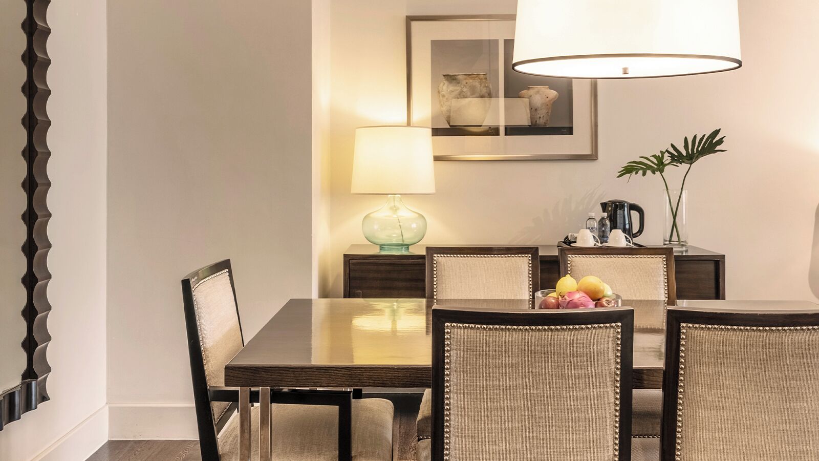 A dining area with a wooden table, four upholstered chairs, a glass-top table, a lamp, a framed picture, and a plant.