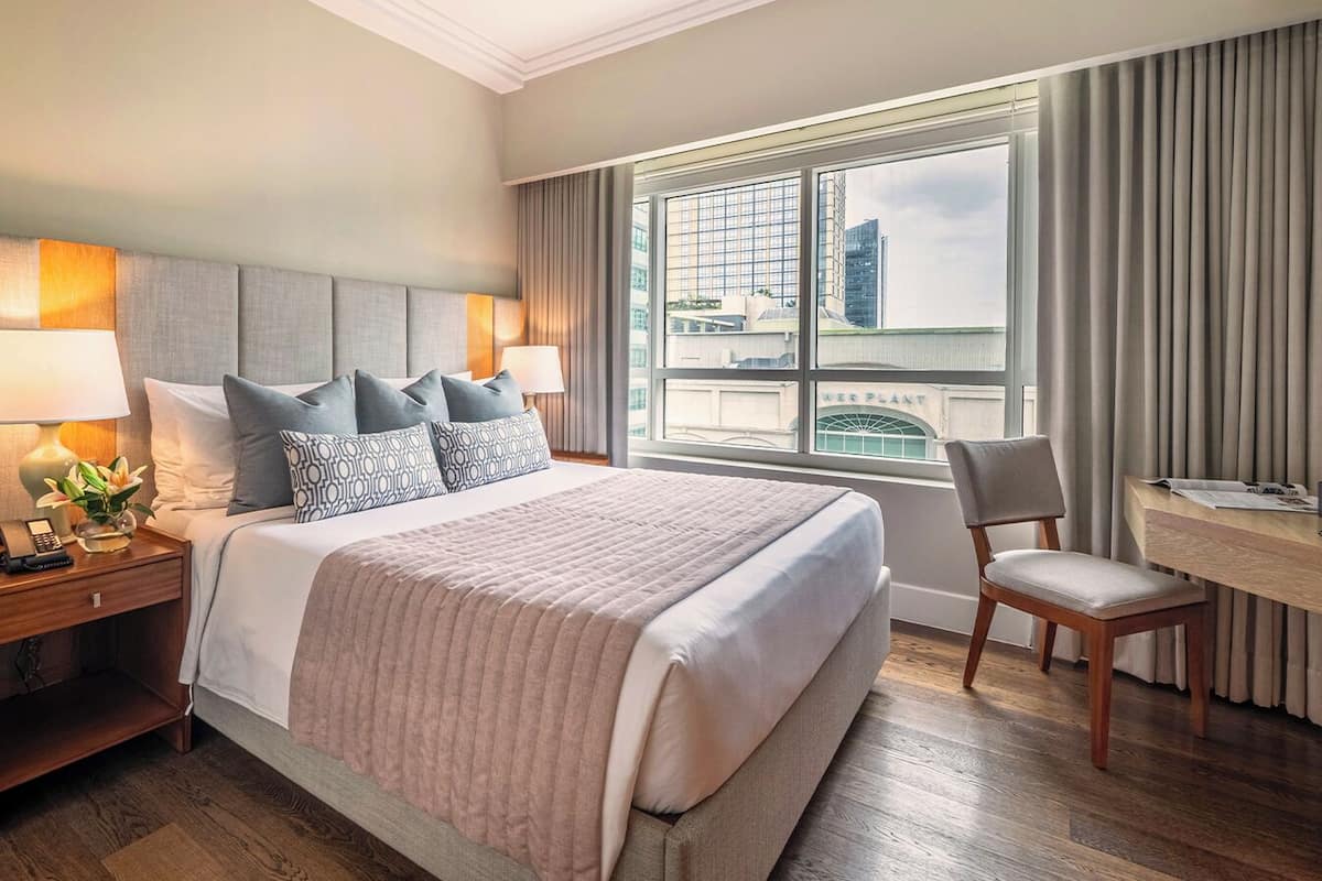 Habitación de hotel con una cama grande, un escritorio, una silla y vistas a un paisaje urbano a través de la ventana.