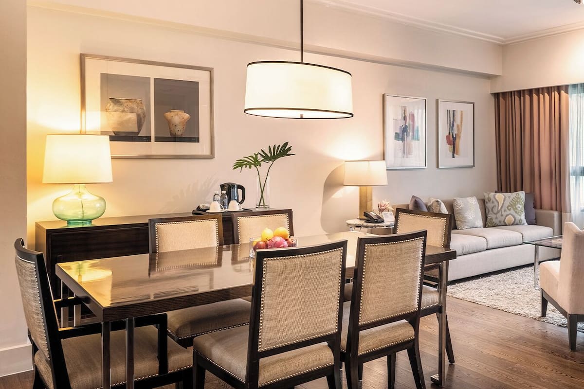 Una sala moderna con comedor, que incluye una mesa de madera, sillas, un sofá y una mesa de centro. Hay dos lámparas fijadas a la pared, una planta en maceta y cuadros en las paredes.