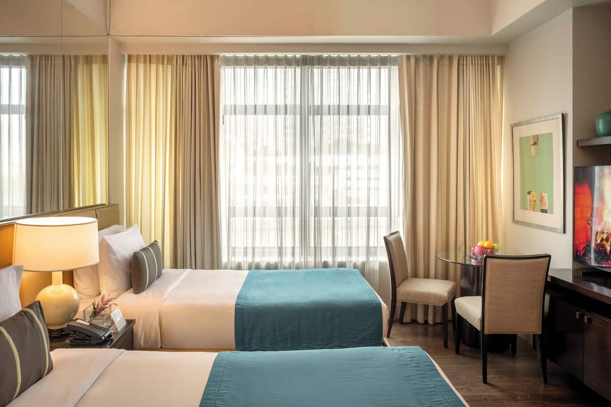 Habitación de hotel con dos camas, un escritorio, una silla, una lámpara y un ventanal grande con cortinas.