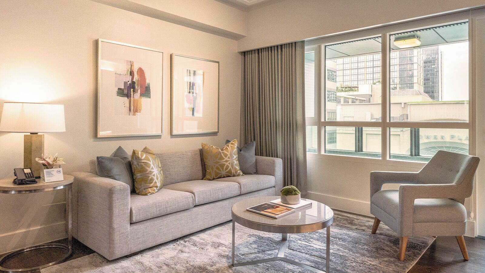 A modern living room with a grey sofa, a round coffee table, and a grey armchair. There are two framed artworks on the wall, a lamp on a side table, and a view of buildings through large windows.