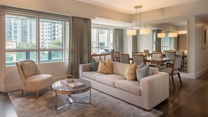 Un salon moderne avec un canapé, un fauteuil et une table basse. On y trouve également un coin repas avec des chaises et une table. De grandes fenêtres offrent une vue sur les bâtiments environnants.