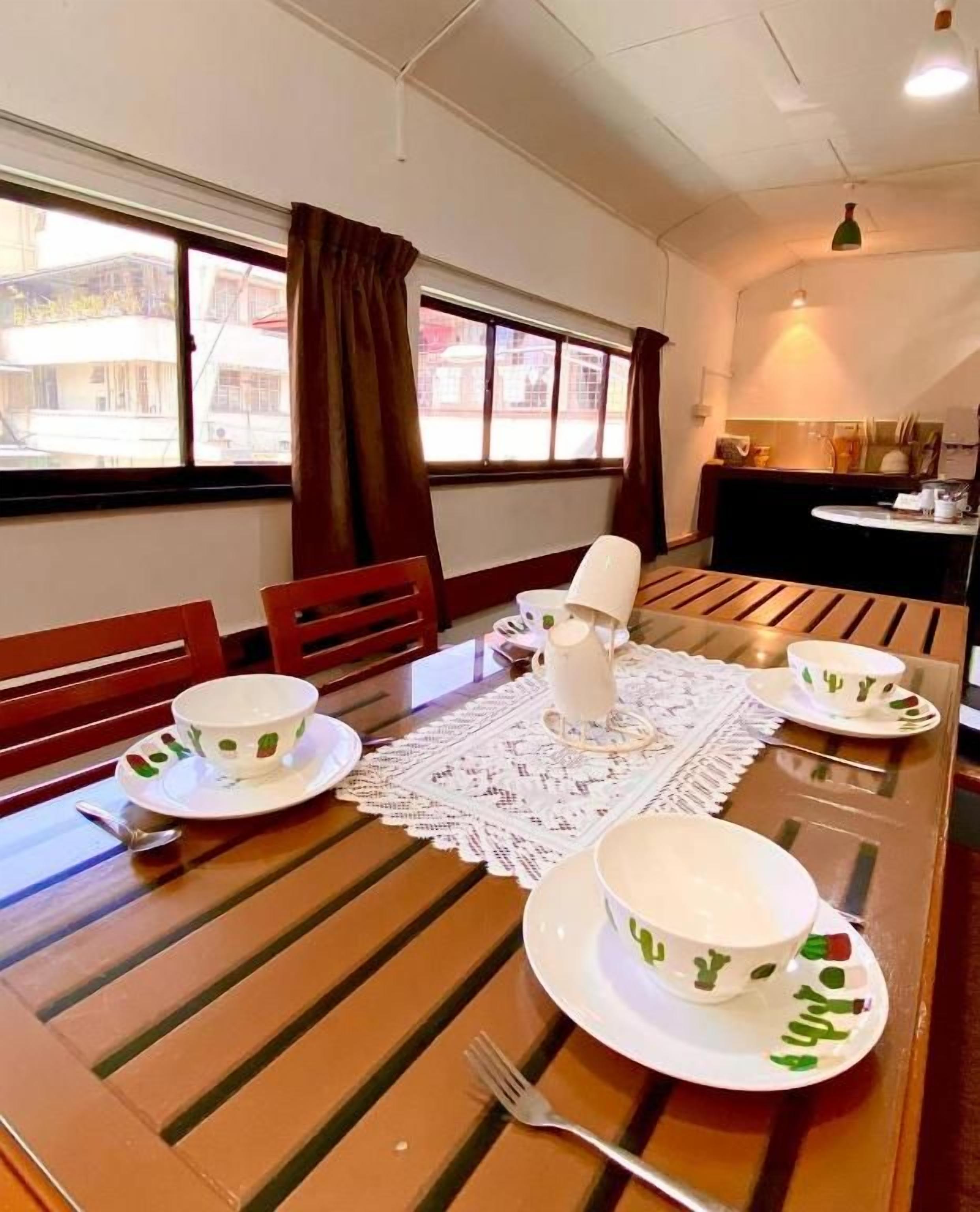 A dining area with a wooden table set for four, a kitchen in the background, and a view of buildings outside.