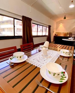 A dining area with a wooden table set for four, a kitchen in the background, and a view of buildings outside. - X-Plorer Backpackers - Hostel (Kota Kinabalu)