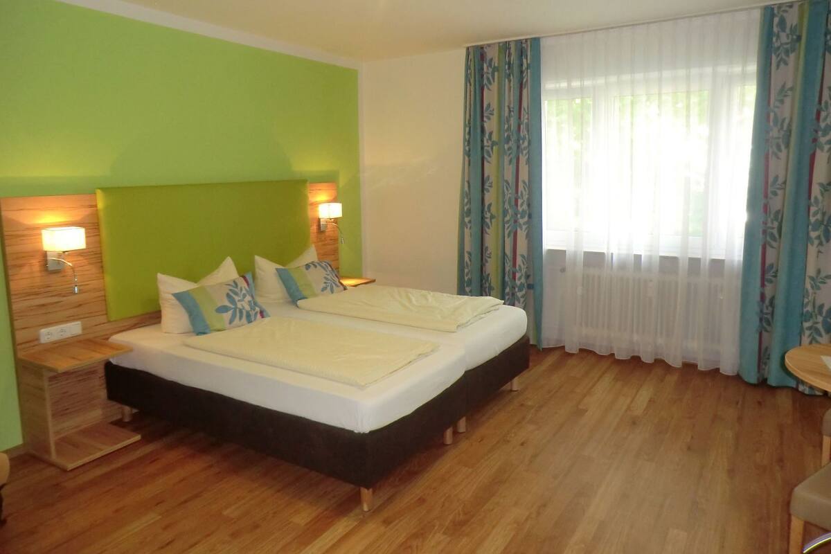 A bedroom with a bed, two bedside lamps, a wooden nightstand, and a window with curtains.