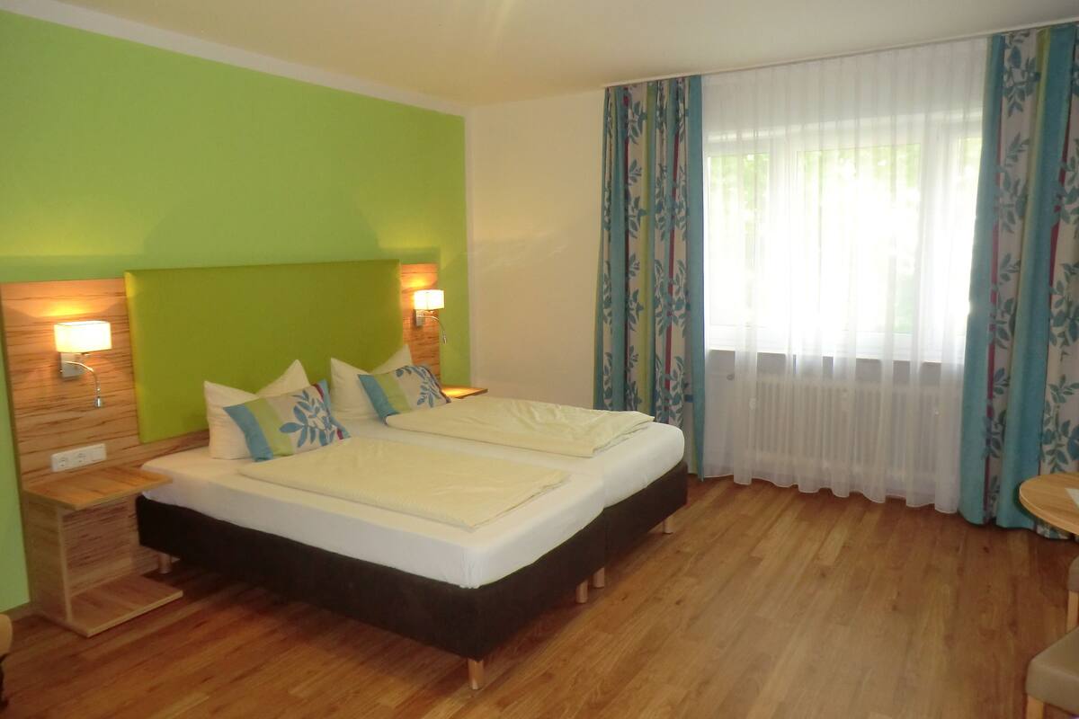 A hotel room with a large bed, two bedside lamps, a wooden nightstand, and a window with floral curtains.