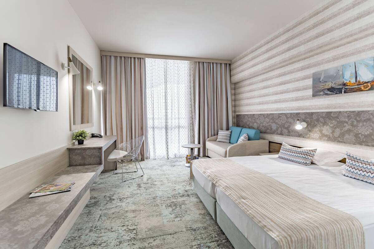 A modern hotel room with a large bed, a sofa, a desk, and a chair. There is a large window with curtains, a painting on the wall, and a small table with a lamp.