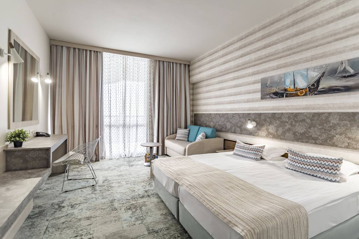 A modern hotel room with a large bed, a desk with a chair, a small table, and a large window with curtains.