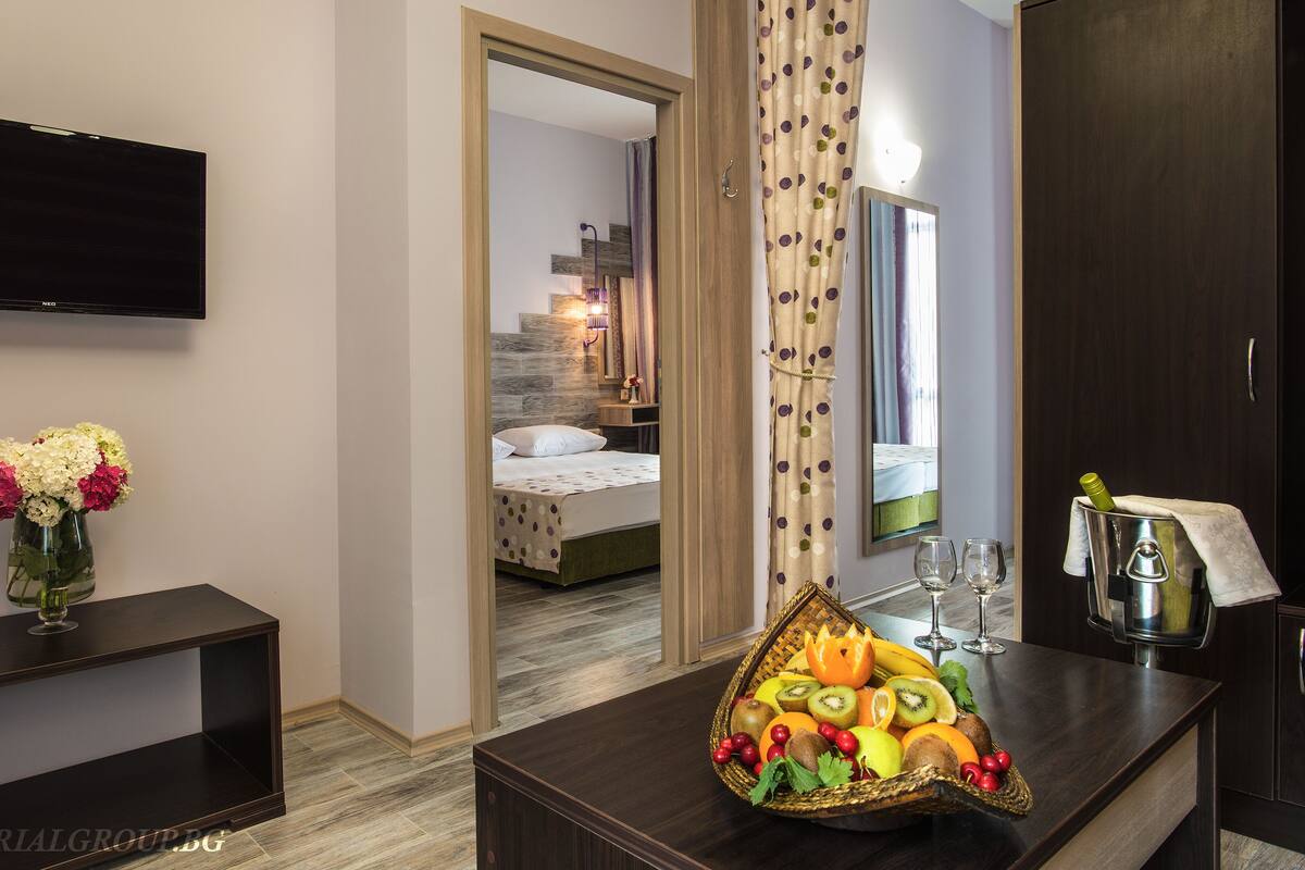 A hotel room with a bed, a TV, a desk with a vase of flowers, and a fruit basket.