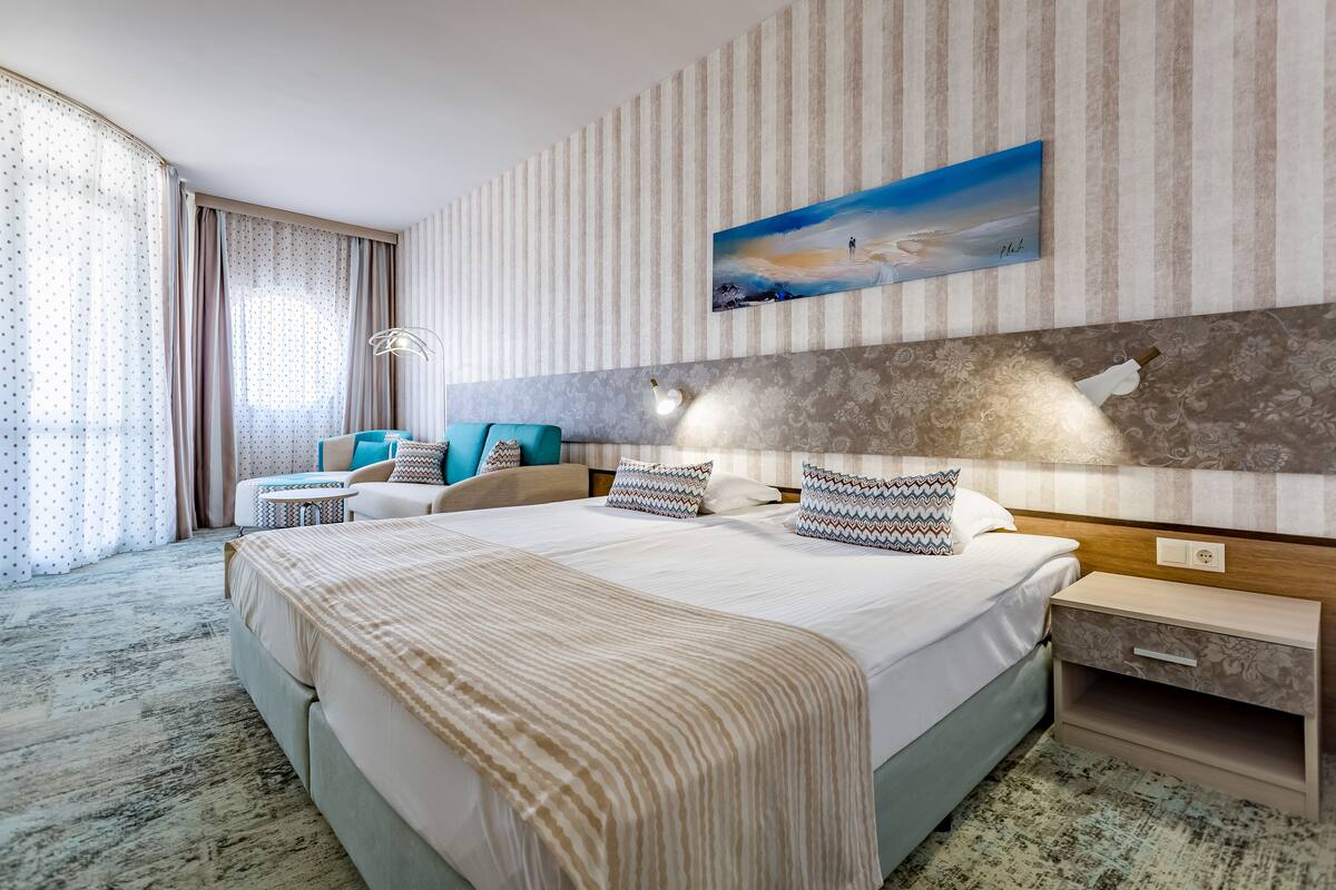 A hotel room with a large bed, a bedside table, a nightstand, a lamp, and a painting on the wall.
