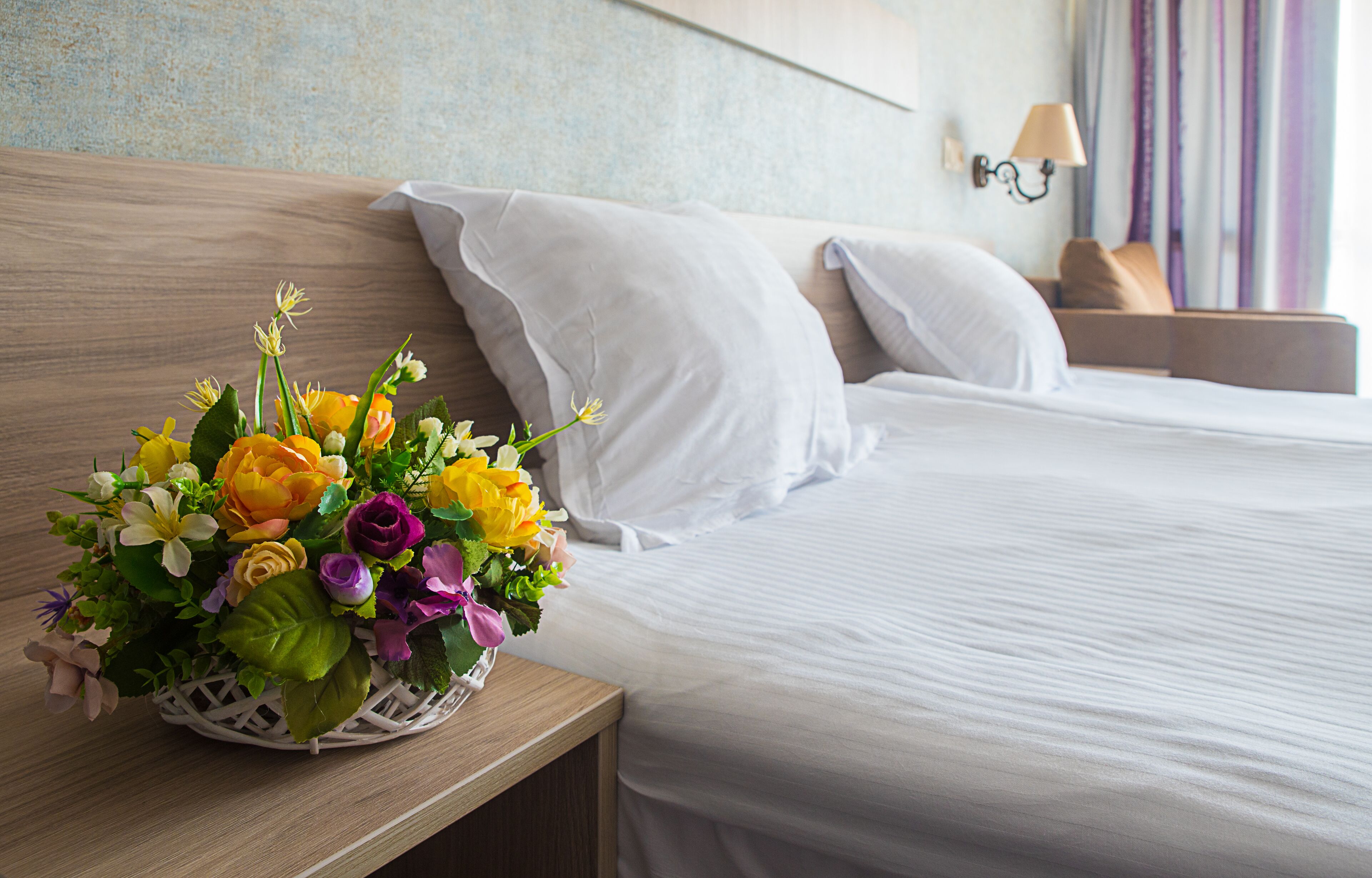 A neatly made bed with white linens, a wooden headboard, and a nightstand with a white lamp and a floral arrangement.