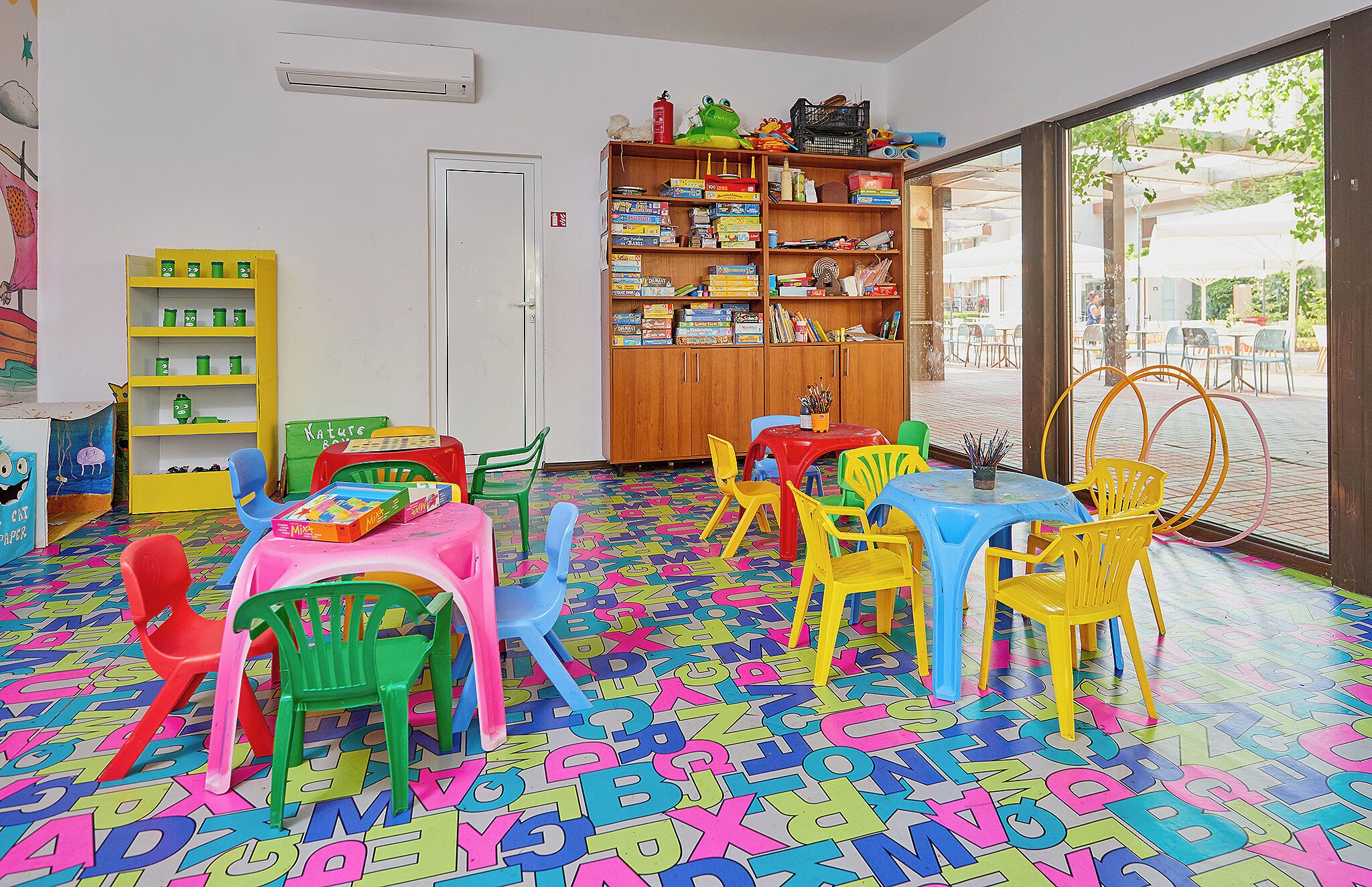 A brightly colored children's playroom with alphabet letters on the floor, a bookshelf, and a table with chairs.