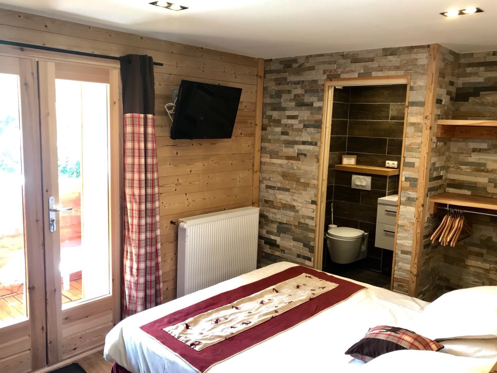 A bedroom with a bed, a window with curtains, a television mounted on the wall, and a bathroom with a toilet and shower.