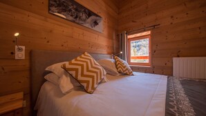 A wooden cabin bedroom with a bed, two pillows, a nightstand, a wall-mounted lamp, a window with a view, and a radiator. - Hôtel Les Escondus (Vars)