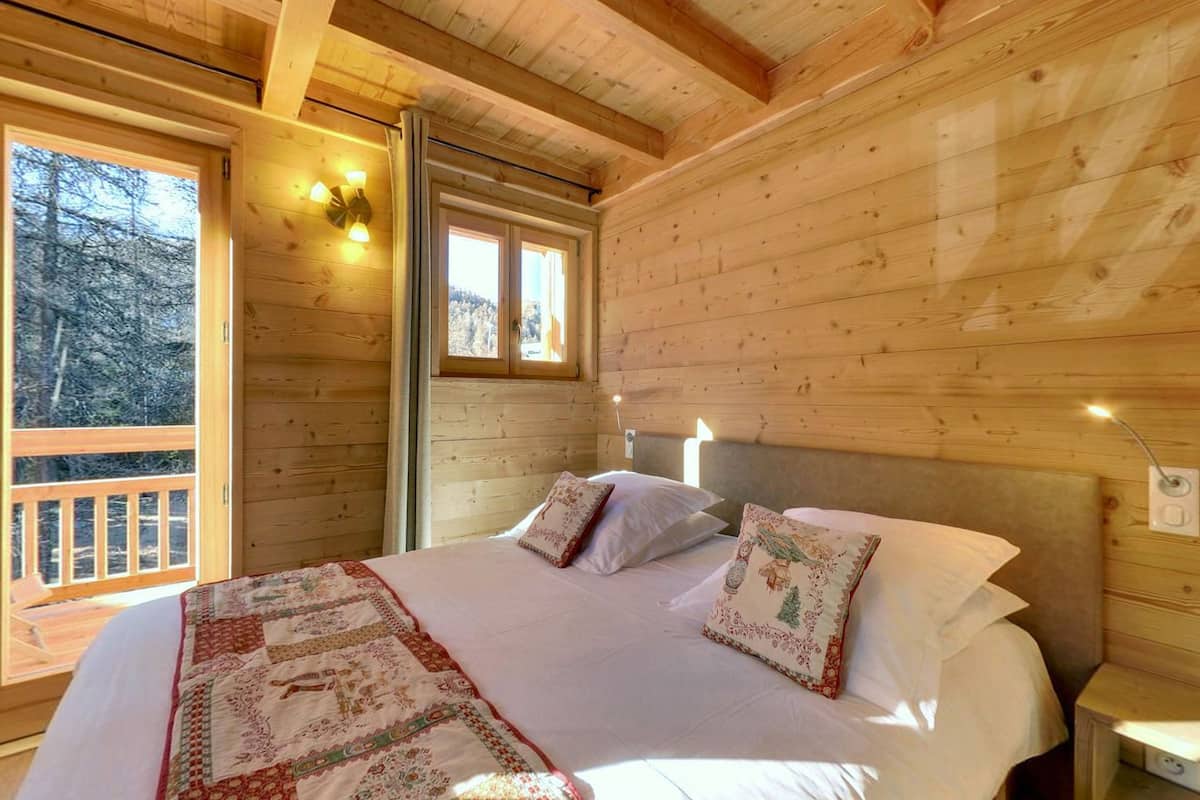 A wooden cabin bedroom with a large bed, two bedside lamps, and a window overlooking a forest.