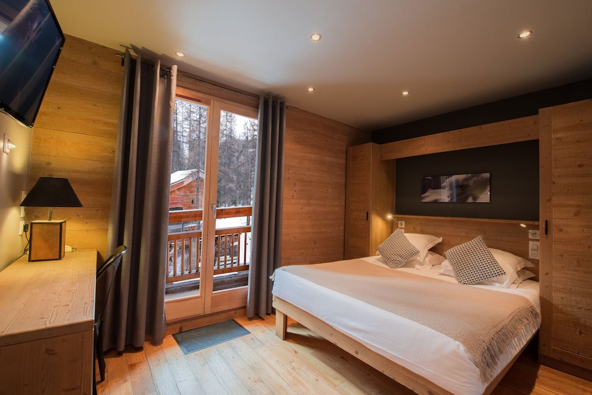 A bedroom with a wooden floor, a bed with white bedding, a desk with a lamp, a TV mounted on the wall, and a window with curtains.