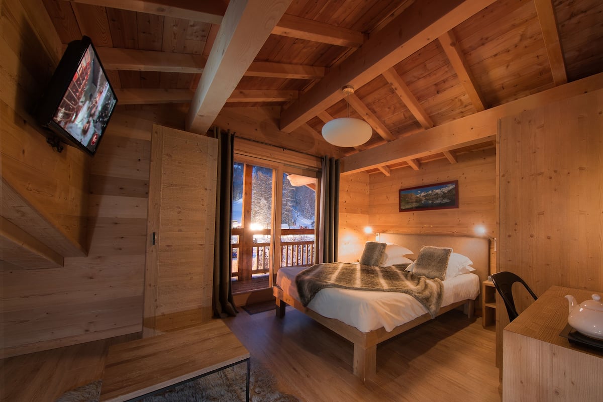 A cozy wooden cabin bedroom with a bed, a television mounted on the wall, and a window overlooking a snowy landscape.