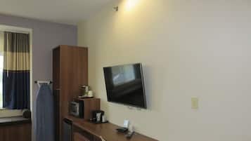 A hotel room with a wooden desk, a flat-screen TV mounted on the wall, a microwave, a coffee maker, and a window with curtains.