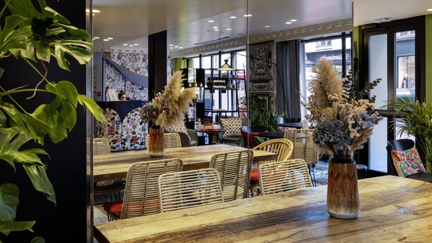 Ein Restaurant mit Holztisch, Korbstühlen, einer großen Vase mit Trockenblumen und einer modernen Deckenleuchte.