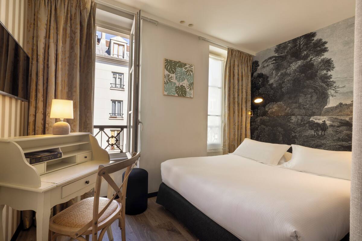 A hotel room with a bed, desk, chair, and a painting on the wall.