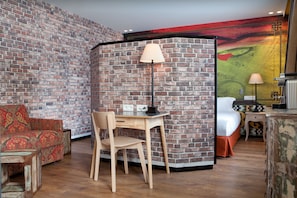 A room with a brick wall, a wooden desk with a lamp, a red patterned sofa, and a bed with a white headboard.