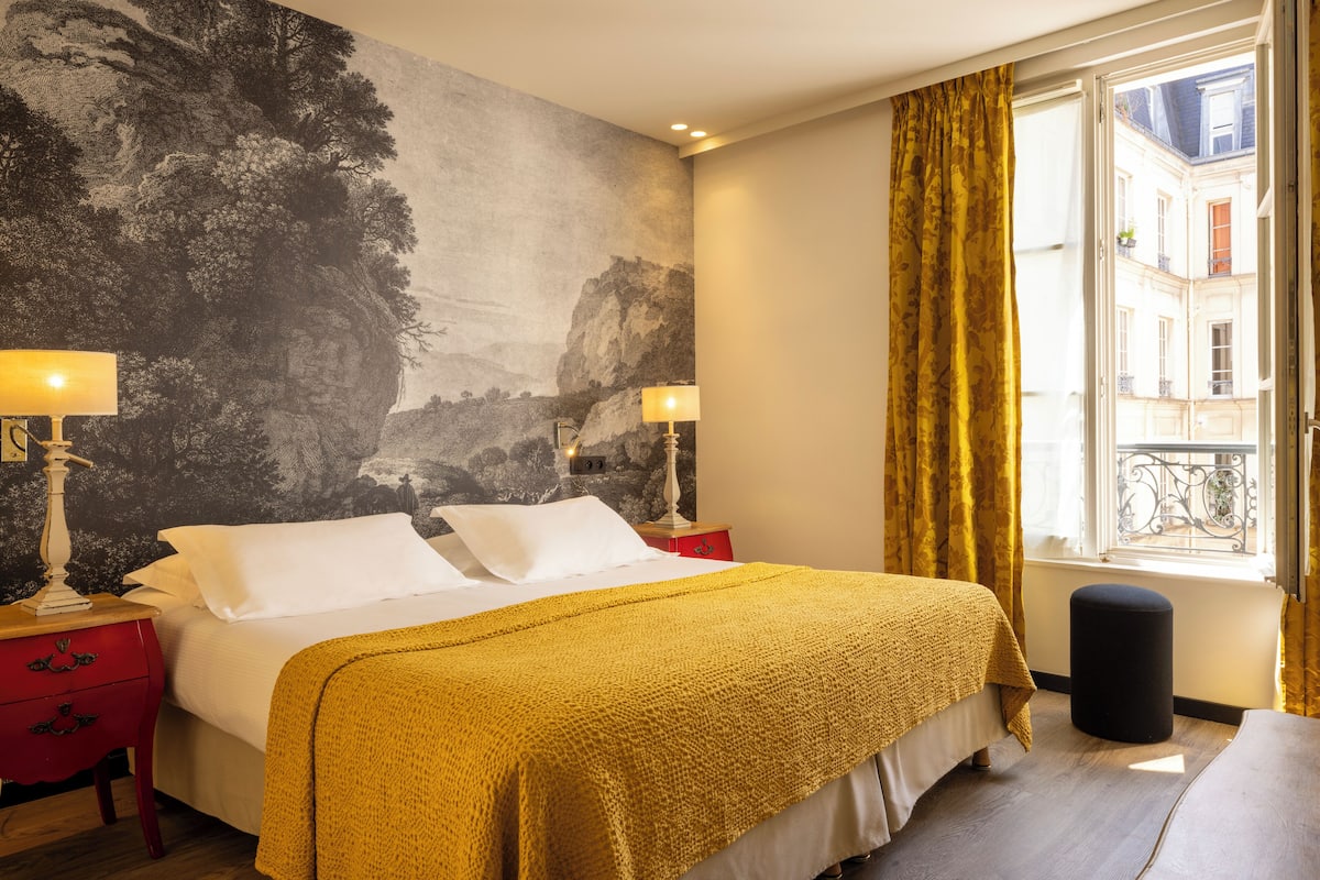 A hotel room with a large bed, two bedside tables with lamps, a window with curtains, and a mural on the wall.