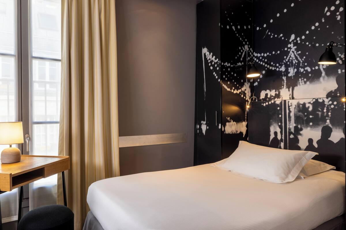 A hotel room with a bed, a desk with a lamp, and a wall with string lights.