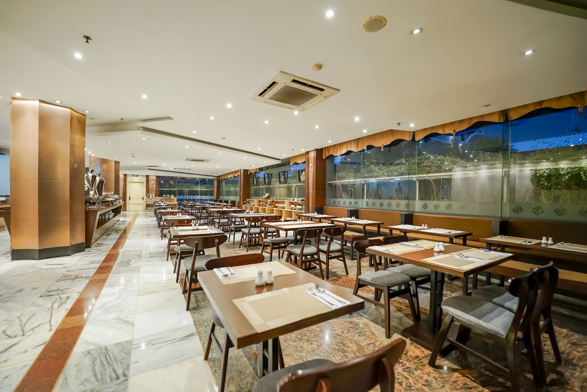 A spacious restaurant with marble flooring, wooden tables and chairs, and large windows offering a view of the outdoors.