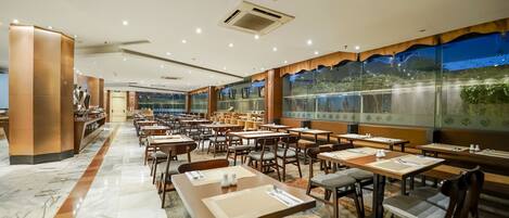 A spacious restaurant with marble flooring, wooden tables and chairs, and large windows offering a view of the outdoors.