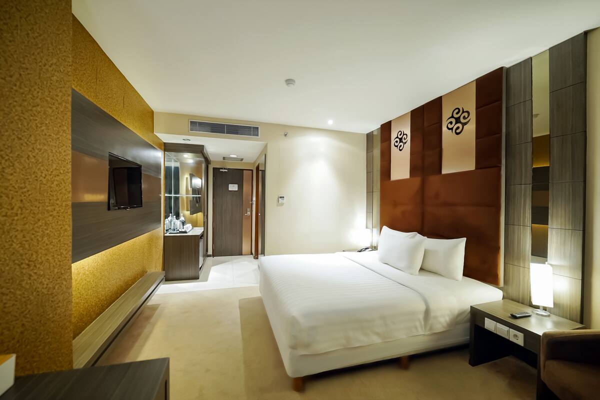 Une chambre d’hôtel avec un grand lit, une table de chevet, un téléviseur fixé au mur et une salle de bain visible à travers une porte ouverte.
