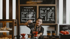 Een barista maakt een drankje in een koffiebar, met een menubord op de achtergrond waarop verschillende koffie- en drankjes staan.