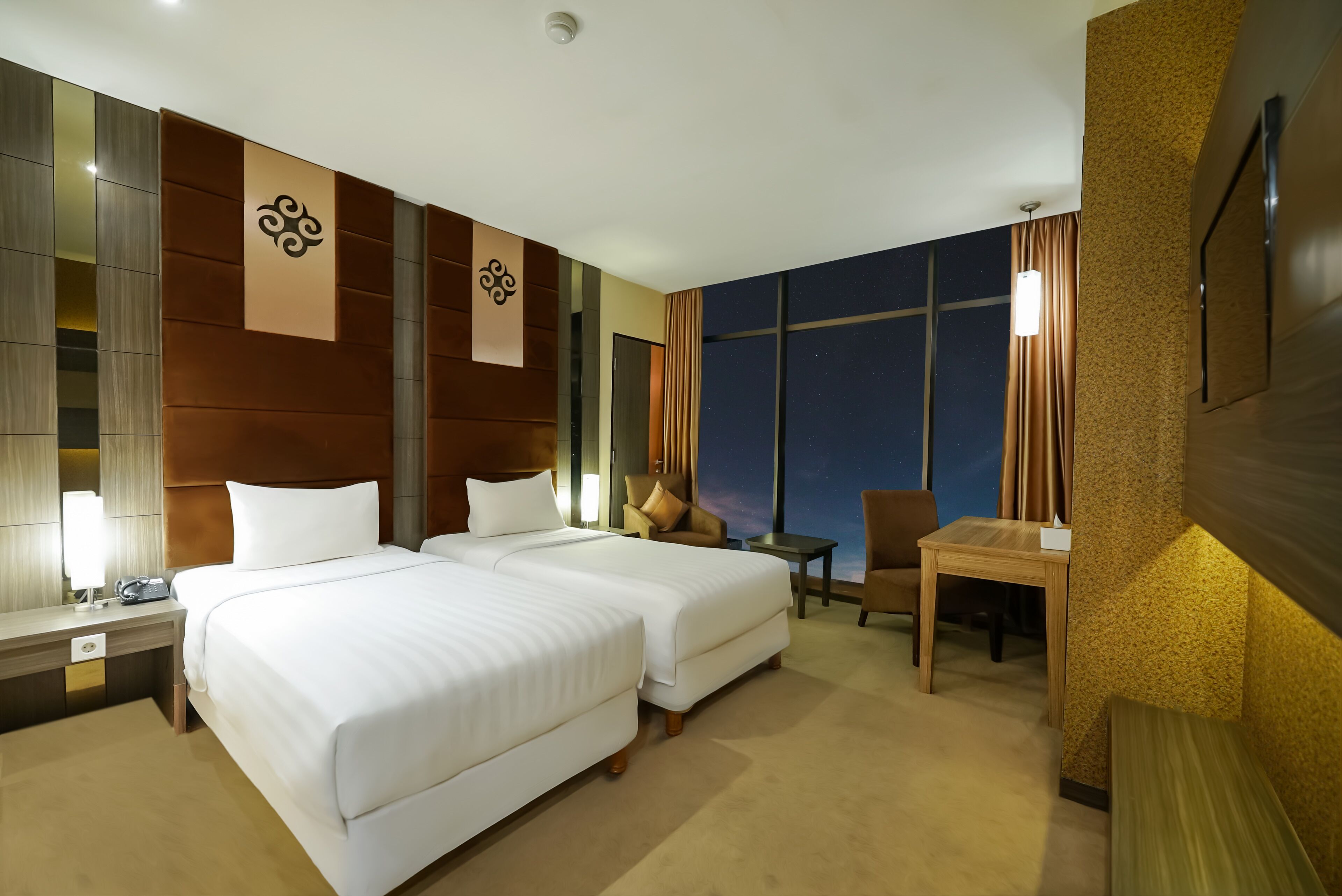 A hotel room with two beds, a desk, a chair, and a large window with curtains.