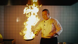 Een kok die een flambé bereidt met een pan boven een tafel in een keuken.