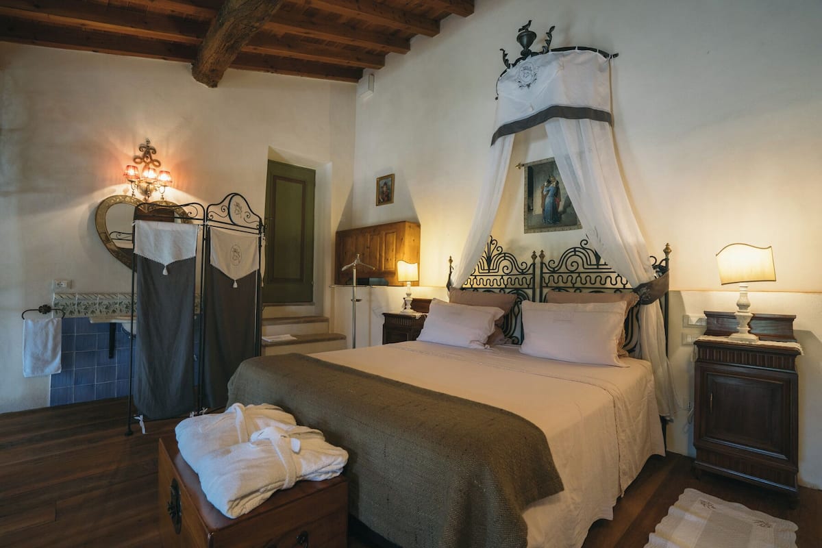 Une chambre à coucher avec un lit à baldaquin, une table de chevet avec une lampe, un banc en bois avec des serviettes et un miroir muni d’un porte-serviettes.