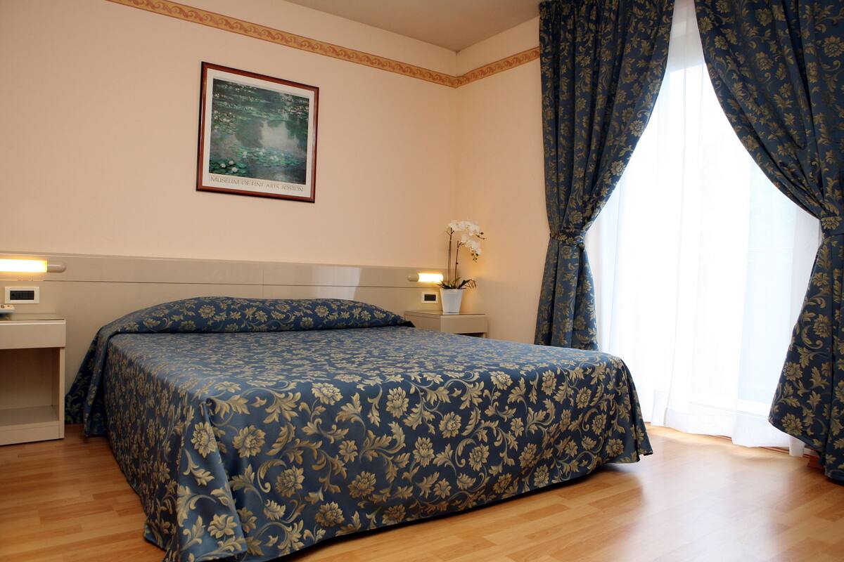 Un lit bien fait, recouvert d’une couvre-lit à motifs floraux, une table de chevet avec une lampe, un tableau encadré au mur et une fenêtre avec des rideaux.