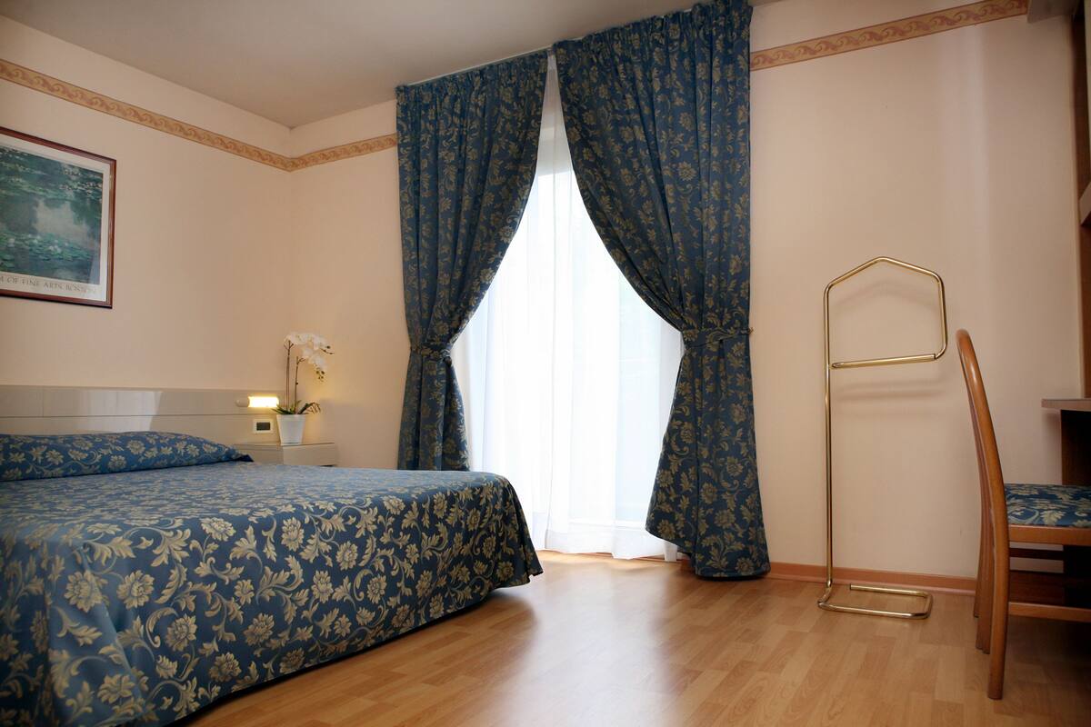 Une chambre d’hôtel avec un lit, une chaise, une table de chevet avec une lampe et une fenêtre avec des rideaux.
