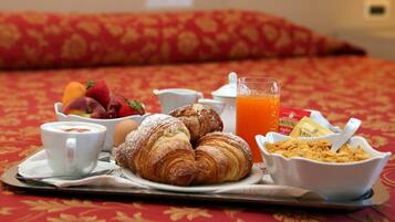 A breakfast tray with croissants, fruit, a cup of coffee, a glass of orange juice, and cereal on a bed with a red patterned bedspread.