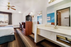 Habitación de hotel con cama, escritorio con televisor, baño con lavamanos y espejo, y ventilador de techo.