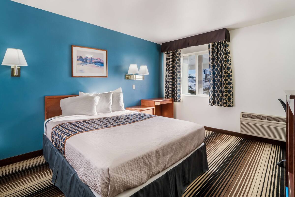 Une chambre d’hôtel avec un grand lit, une table de chevet, deux lampes fixées au mur, un tableau encadré, des rideaux à motifs et une fenêtre donnant sur un beau paysage.