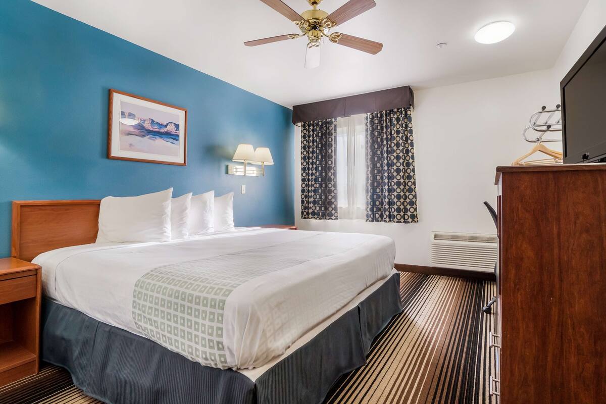 Une chambre d’hôtel avec un grand lit, un ventilateur de plafond, un tableau encadré au mur, un bureau avec une lampe et une commode.