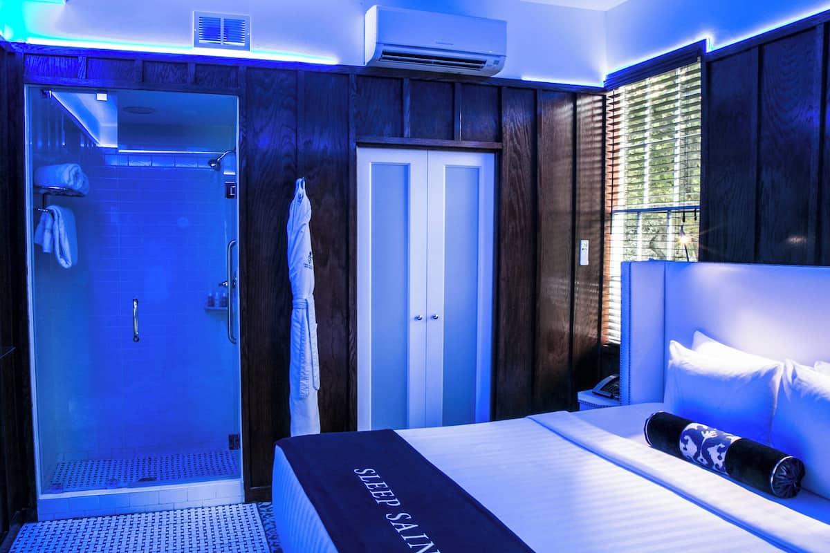 A modern hotel room with a glass-enclosed shower, a bed with a 'Sleep Saint' blanket, and a view of the outdoors through a window.