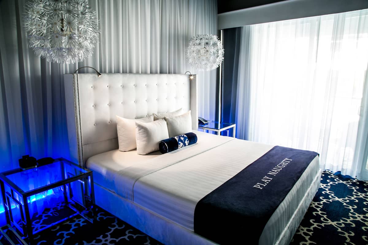 A large bed with a tufted headboard, multiple pillows, and a blanket with "PLAY NIGHT" text. There is a glass side table with a blue light, and a patterned rug on the floor.