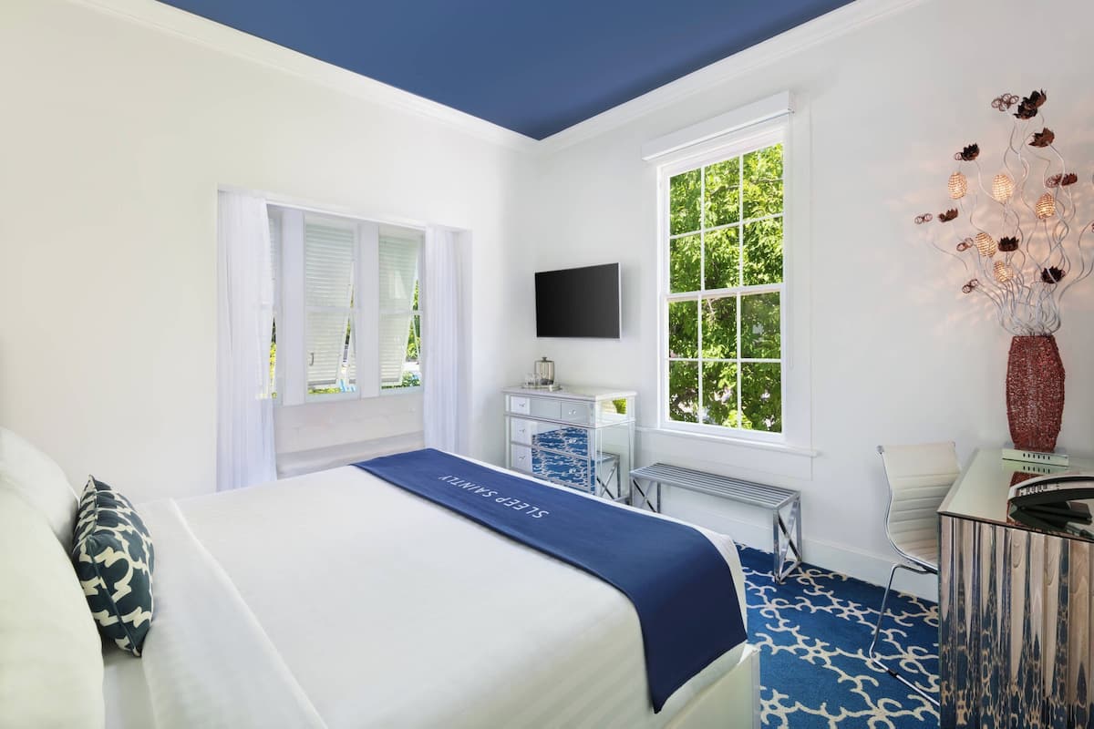 A hotel room with a bed, a desk, a chair, a TV, and a window with a view of greenery.